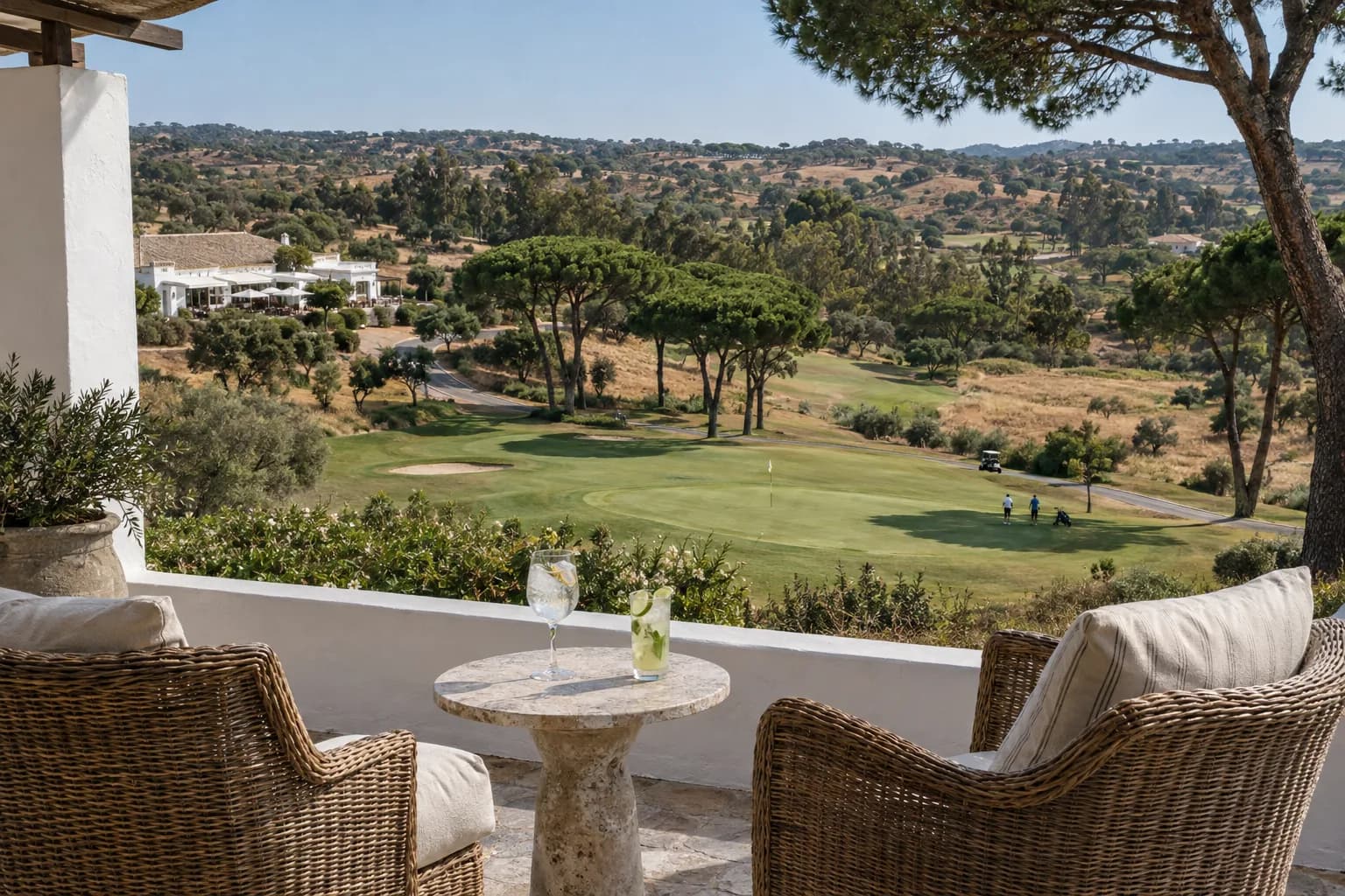 A clubhouse terrace with two drinks on a stone table, the course and pines below